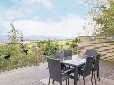 An outdoor terrace with a table and six chairs overlooking a landscape with hills and trees at Hilltop Lodge in Corwen
