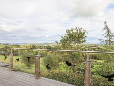 A balcony with wooden posts and glass railing overlooking a green landscape with trees and hills at Hilltop Lodge in Corwen