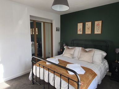 A bedroom with a metal bed frame, white bedding, two beige pillows, folded white towels, a brown throw, bedside tables with lamps, three framed pictures on a dark green wall and mirrored closet doors at No.37 in Beverley