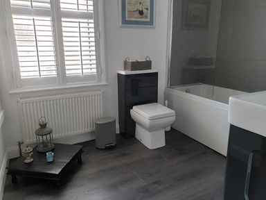 A bathroom with a toilet, bathtub, sink, window with shutters, radiator, small table with decorations, and a trash bin at No.37 in Beverley