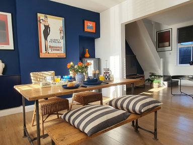 A dining area with a wooden table, wicker chairs, striped cushions on a bench, and framed posters on blue and white walls at No 37 in Beverley
