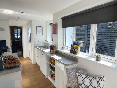 A kitchen with wooden floor white cabinets a countertop with a cutting board bread and croissants a sink books and plants on the windowsill and cushions under the window at No 37 in Beverley