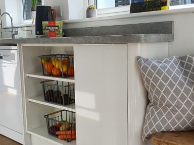 A kitchen countertop with a box of Yorkshire Tea and a kettle above shelves with baskets of fruit and vegetables next to a window with a pillow on a bench at No.37 in Beverley