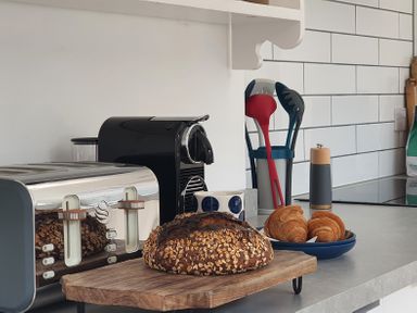 A kitchen countertop with a loaf of bread on a wooden board plate of croissants a toaster coffee machine and utensils in a container at No.37 in Beverley