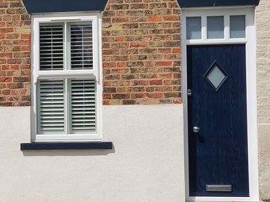 A brick wall with a white window with shutters and a dark blue door with a diamond shaped window at No 37 in Beverley