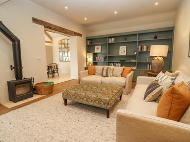 A living room with two sofas a patterned ottoman a wood burning stove and a built in bookshelf at Hawthorn Barn in Northleach