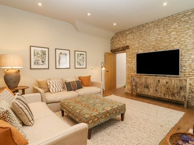 A living room with two beige sofas a patterned ottoman lamp and a large television on a rustic wooden cabinet at Hawthorn Barn in Northleach