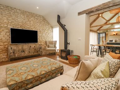 A living room with a patterned ottoman a beige sofa a wood stove and a dining area in the background at Hawthorn Barn in Northleach