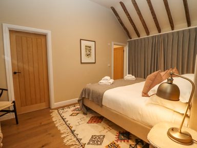 A bedroom with a bed pillows and blankets a bedside lamp a patterned rug wooden doors and ceiling beams at Hawthorn Barn in Northleach