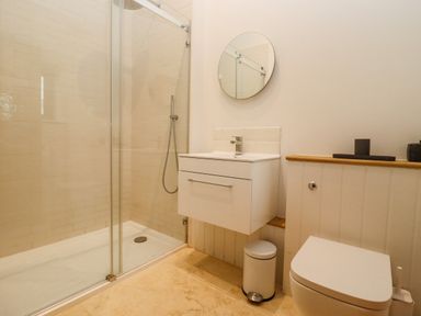 A bathroom with a shower area sink with cabinet round mirror toilet and small trash can at Hawthorn Barn in Northleach