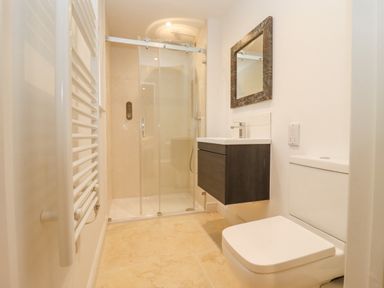 A bathroom with a shower, a wall-mounted sink, a mirror, a toilet, and a towel radiator at Hawthorn Barn in Northleach