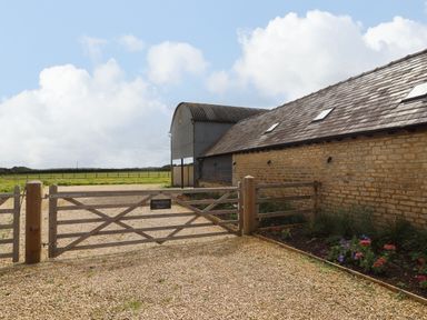 A gravel driveway with a wooden gate and a stone barn next to a flower bed at Hawthorn Barn in Northleach
