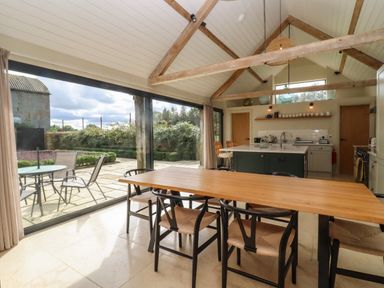 A dining area with a wooden table and chairs next to a kitchen with an island and large glass doors overlooking a patio at Hawthorn Barn in Northleach