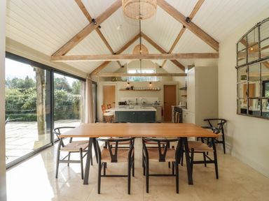 A dining area with a wooden table and chairs next to a kitchen island under a vaulted ceiling with wooden beams at Hawthorn Barn in Northleach