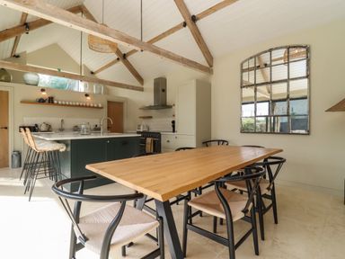 A dining area with a wooden table and black chairs next to a kitchen island with stools and exposed wooden beams at Hawthorn Barn in Northleach
