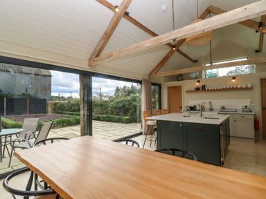 A kitchen with a wooden dining table black chairs an island with bar stools glass sliding doors and exposed wooden beams at Hawthorn Barn in Northleach