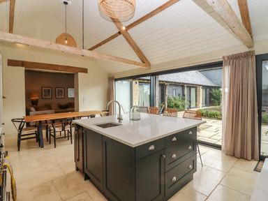 A kitchen with an island and sink adjacent to a dining area with a wooden table and chairs at Hawthorn Barn in Northleach