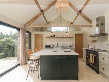 A kitchen with an island, bar stools, large windows, wooden beams, and appliances at Hawthorn Barn in Northleach