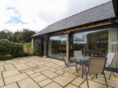 An outdoor patio area with a round glass table and four chairs next to a building with large glass sliding doors and a tiled roof at Hawthorn Barn in Northleach