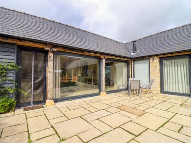 An outdoor patio with stone tiles a round table and four chairs outside a building with large glass windows at Hawthorn Barn in Northleach