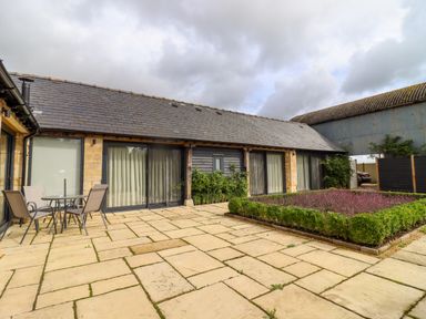 An outdoor patio area with a table and chairs next to a building with large glass doors and a hedge garden at Hawthorn Barn in Northleach