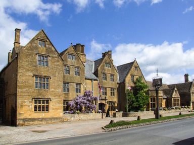 A historic stone building with multiple peaked roofs and chimneys along a street with flags and purple flowers at The Cotswold Holiday Lodge in Evesham