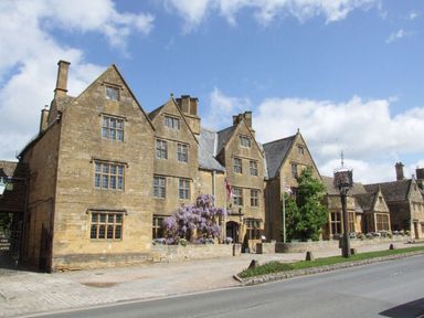 A historic stone building with multiple peaked roofs next to a road with Union Jack flags and flowering plants at The Cotswold Holiday Lodge in Evesham