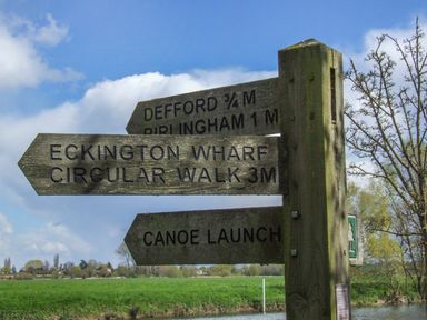 A wooden signpost with direction signs for Defford, Birlingham, Eckington Wharf Circular Walk, and Canoe Launch in a rural area