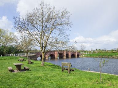 A riverside park with picnic tables benches trees and a stone bridge over the water at The Cotswold Holiday Lodge in Evesham