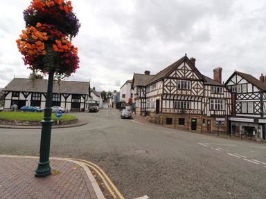 A street with timber-framed buildings a roundabout with green grass and a lamp post with flower baskets in Ruthin