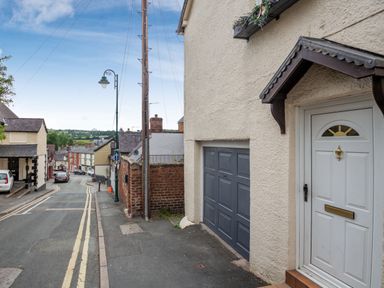 A street with a white door and garage on a beige building with houses and cars along the road at Dove Cottage in Ruthin