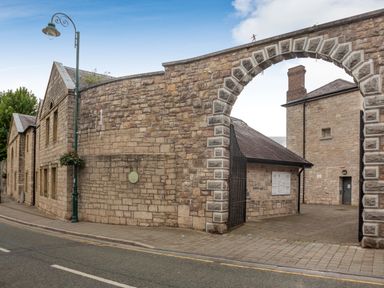 A stone archway leading to stone buildings with a lamp post and sidewalk in front at Dove Cottage in Ruthin