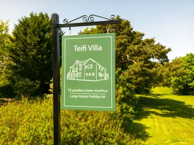 A green sign on a black post with drawing of house and text about a large luxury holiday let surrounded by grass and trees at Teifi Villa in St Dogmaels