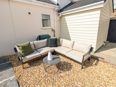 An outdoor seating area with an L-shaped sofa with cushions and a round table on a gravel surface at Teifi Villa in St Dogmaels