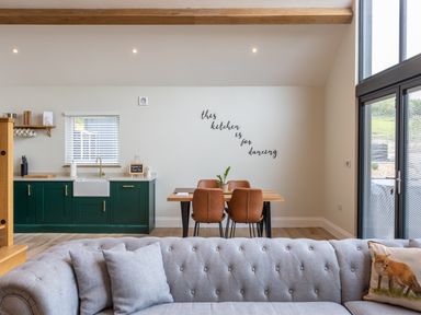 A kitchen area with green cabinets a wooden dining table with four brown chairs and a gray tufted sofa in the foreground at Foxes Den Allerston near Thornton Dale