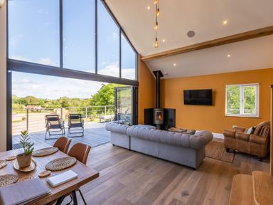 A living room with a gray sofa a wood burning stove and large windows opening to a balcony at Foxes Den Allerston near Thornton Dale