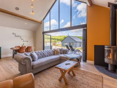 A living room with a gray sectional sofa wooden coffee table and black wood stove next to large windows leading to an outdoor deck at Foxes Den in Allerston near Thornton Dale