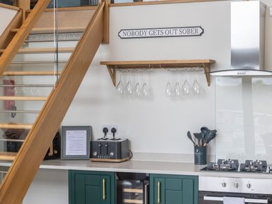 A kitchen area with green cabinets a toaster cooking utensils and glassware hanging under a wooden shelf at Foxes Den Allerston near Thornton Dale