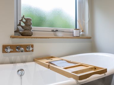 A bathroom with a white bathtub wooden tray on the tub and a window ledge with decorative items at Foxes Den in Allerston near Thornton Dale