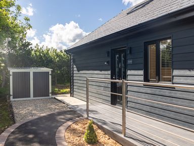 An exterior view of a black wooden building with a metal railing and a small shed on a gravel area at Foxes Den in Allerston near Thornton Dale