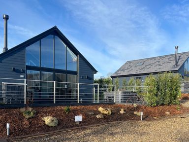 Two modern dark buildings with large glass windows and chimneys surrounded by mulch and shrubs at Foxes Den in Allerston near Thornton Dale