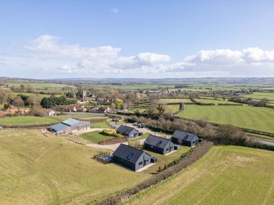 A cluster of black cottages and a large barn in a rural landscape with fields and a village in the distance at Foxes Den in Allerston near Thornton Dale