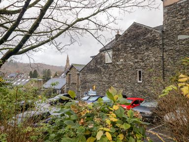 A parking lot with cars next to stone buildings with a tree and plants in the foreground at Dixon Court in Ambleside