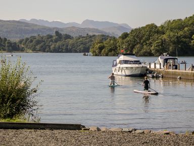 Two people paddleboarding on a lake near boats and a dock with trees and hills in the background at Dixon Court in Ambleside