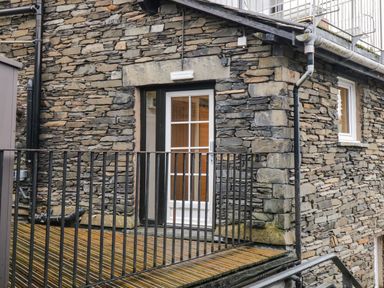 A stone exterior wall with a white-framed glass door and a metal railing on a wooden deck at Dixon Court in Ambleside