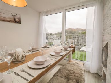 A dining area with a wooden table set with plates bowls glasses and cutlery next to a window overlooking a balcony and hills at Dixon Court in Ambleside