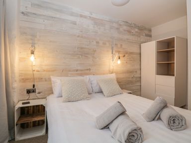A bedroom with a bed with white linens and two beige pillows two rolled towels bedside shelves and lit wall lamps at Dixon Court in Ambleside