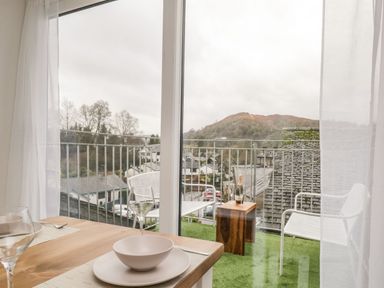 A dining table set with plates and glasses next to a balcony with chairs and a small wooden table overlooking houses and hills at Dixon Court in Ambleside