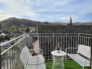A balcony with two white chairs and a small table holding a bottle and glasses overlooking rooftops and hills at Dixon Court in Ambleside