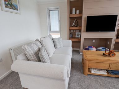 A living room with a light sofa with cushions a wooden coffee table a wall mounted television and built in shelves at Hideaway Lodge in Runswick Bay